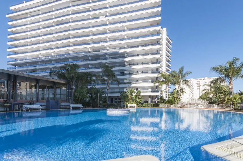 A swimming pool sits beside a modern high-rise building and palm trees.
