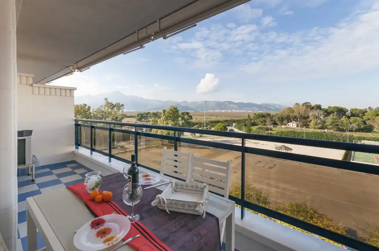 A covered balcony with a table and seating overlooks green fields and distant mountains.