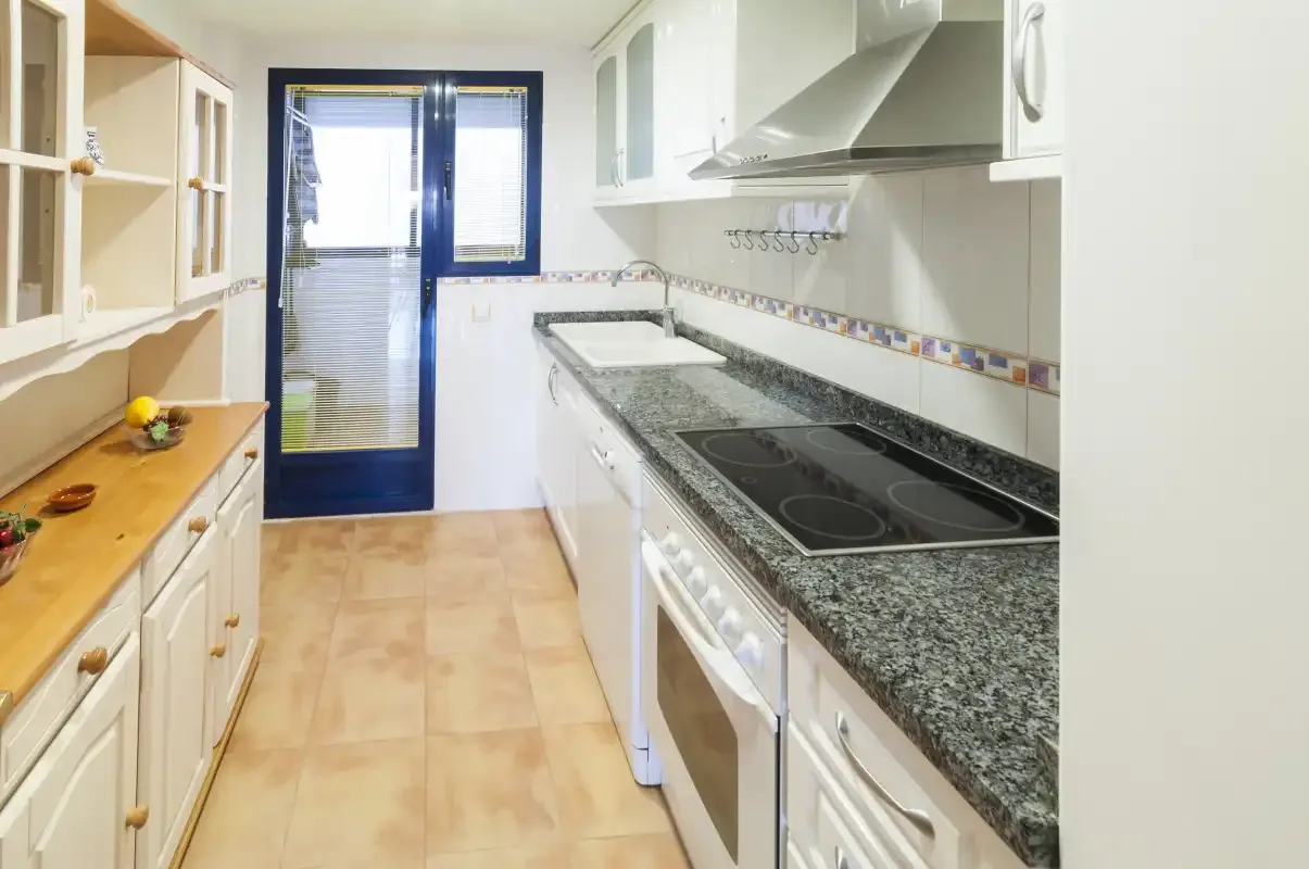 A narrow kitchen with white cabinets, dark countertops, and a tiled floor.