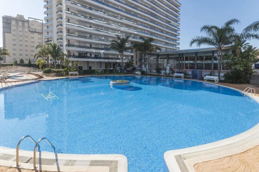 A large outdoor swimming pool sits beside a high-rise building and palm trees.