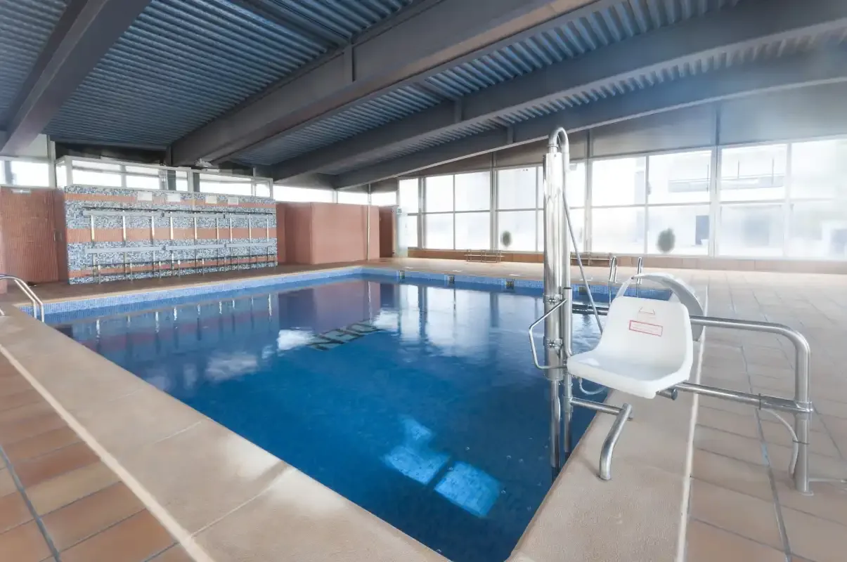 Indoor swimming pool with blue water, a tiled deck, and large windows.