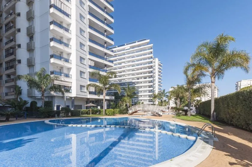 A swimming pool sits in front of modern apartment buildings and palm trees.
