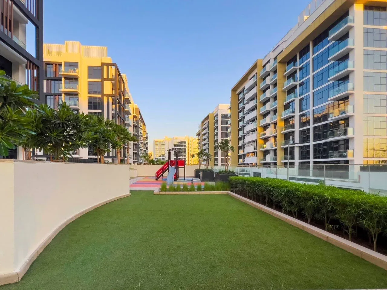 Un patio ajardinado entre edificios de apartamentos modernos, con un pequeño parque infantil al fondo.