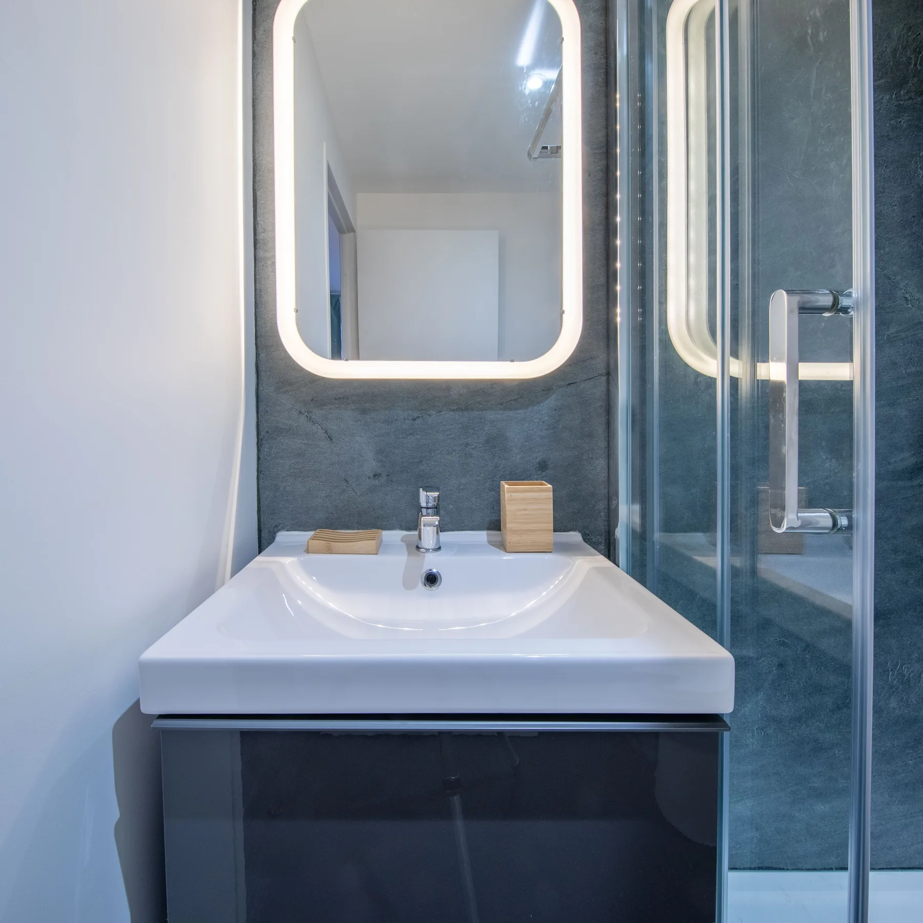 A bathroom vanity with a sink and illuminated mirror beside a glass shower.