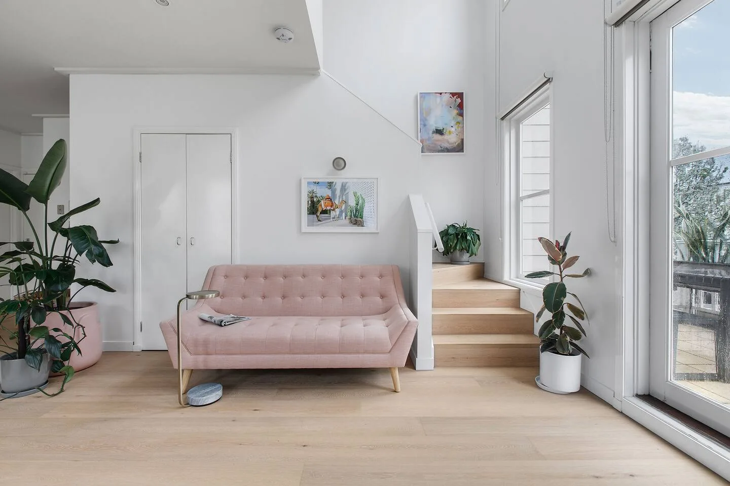 Una sala luminosa con un sofá rosa junto a grandes ventanales y plantas en macetas.