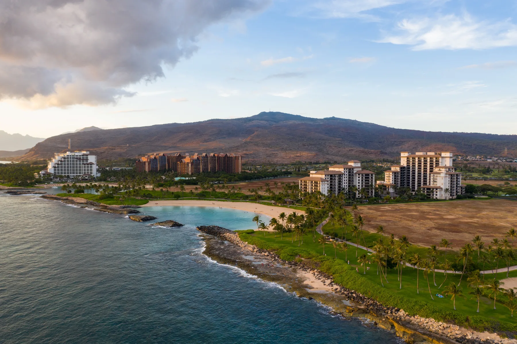 Un complejo turístico costero se encuentra junto a una costa verde, con montañas al fondo.