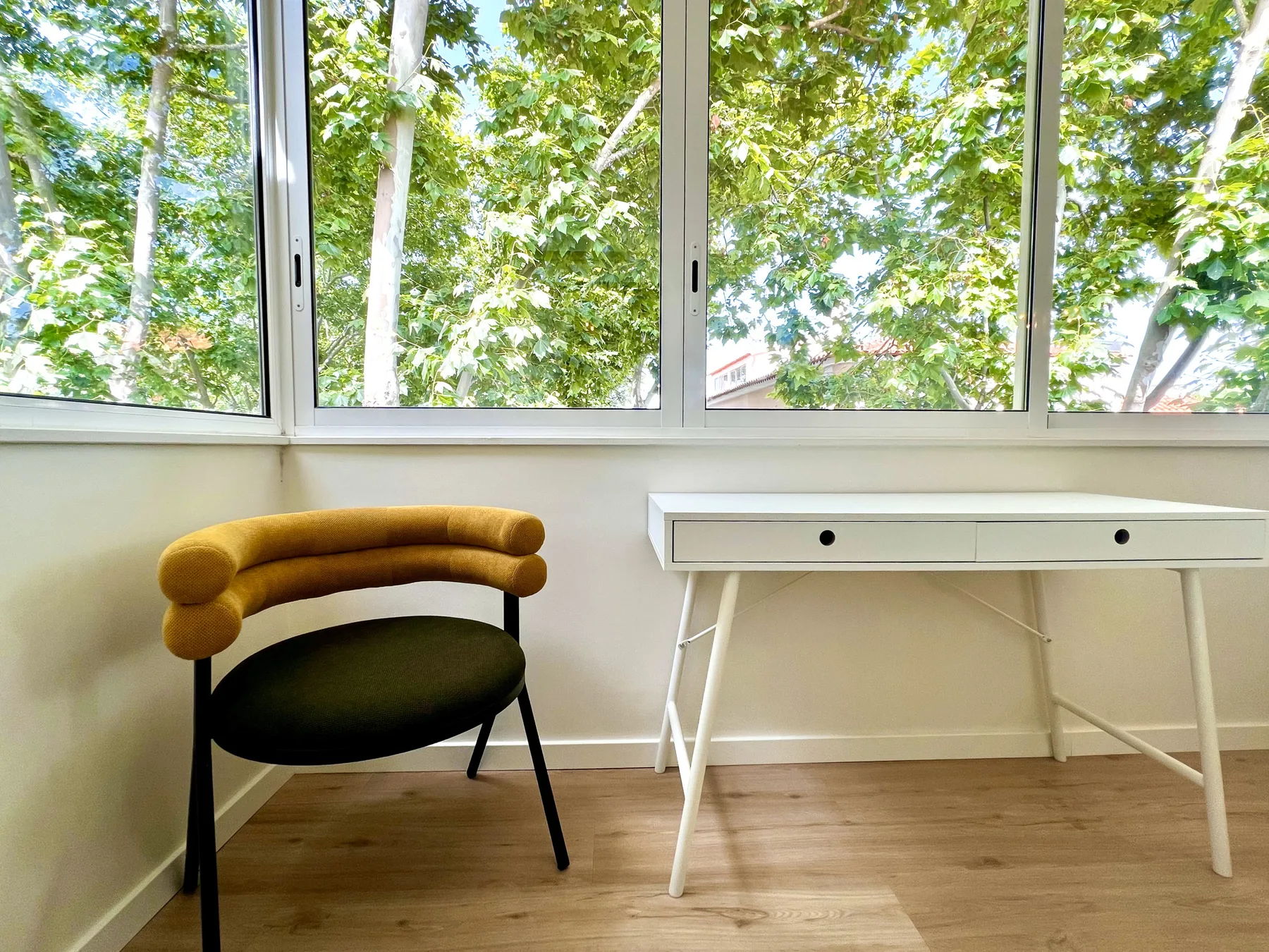 Una habitación luminosa con una silla y un escritorio junto a grandes ventanas con vista a árboles frondosos.