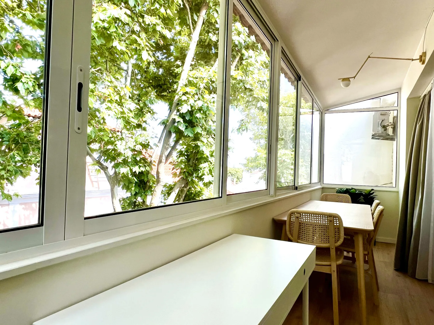Un balcón cerrado y luminoso con una mesa larga junto a ventanas frondosas.