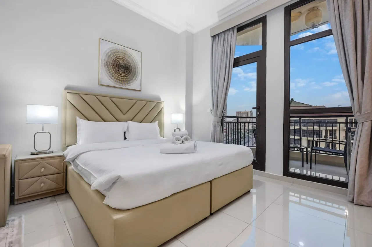 A bright bedroom with a neatly made bed beside tall windows overlooking a balcony.