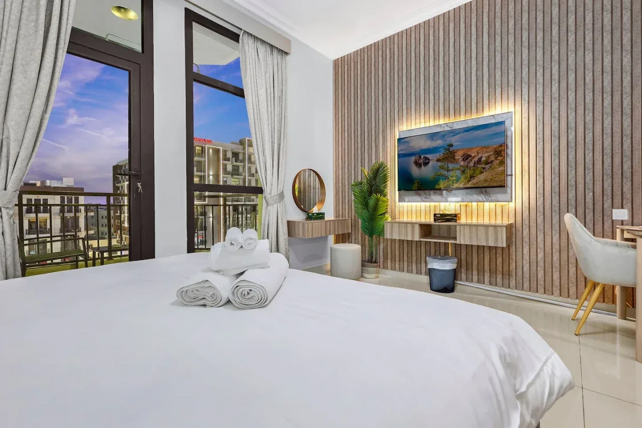 A bright bedroom with a neatly made bed, tall windows, and a wall-mounted television.