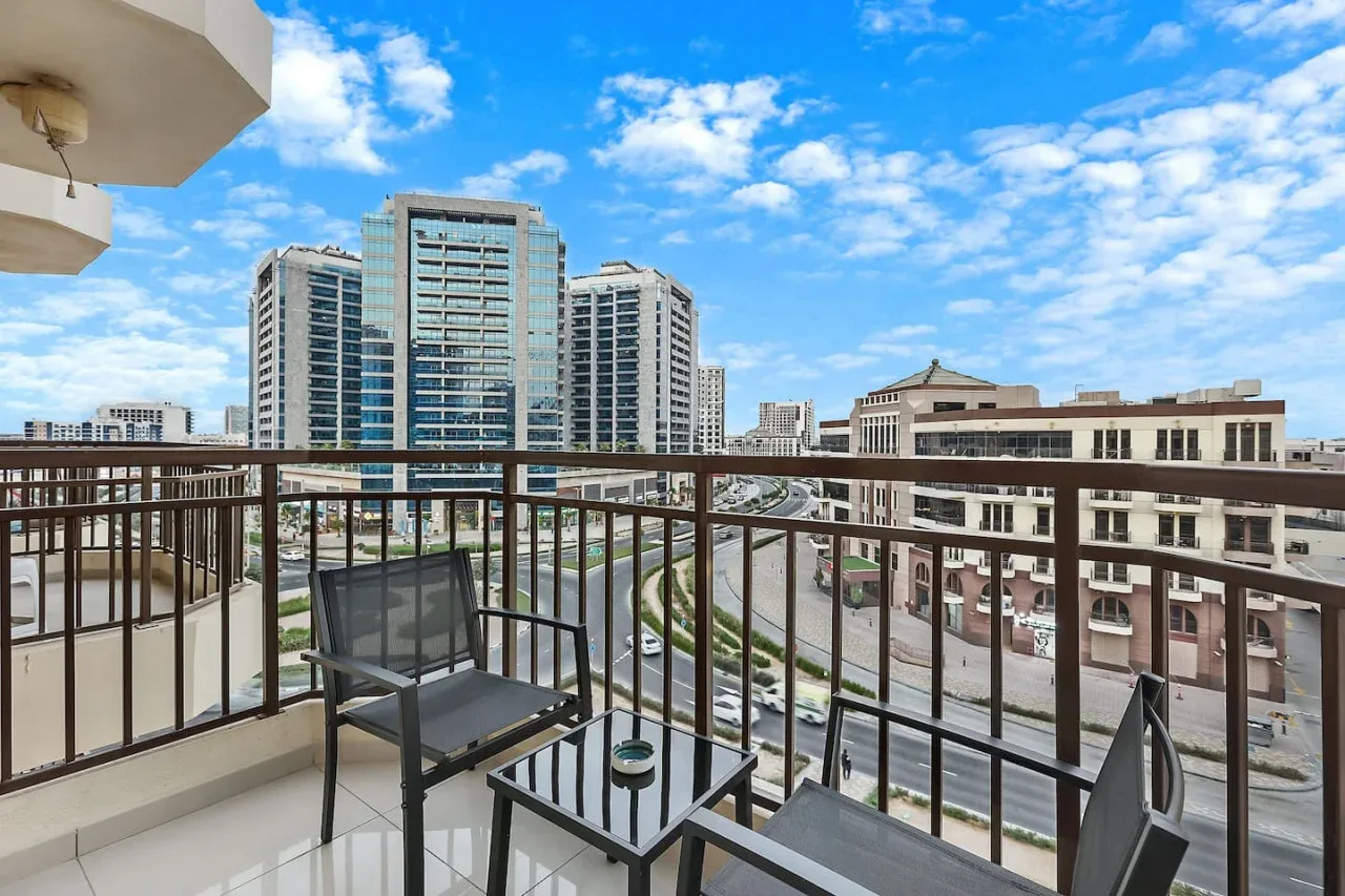 A balcony seating area overlooks nearby buildings beneath a bright, cloud-dotted sky.
