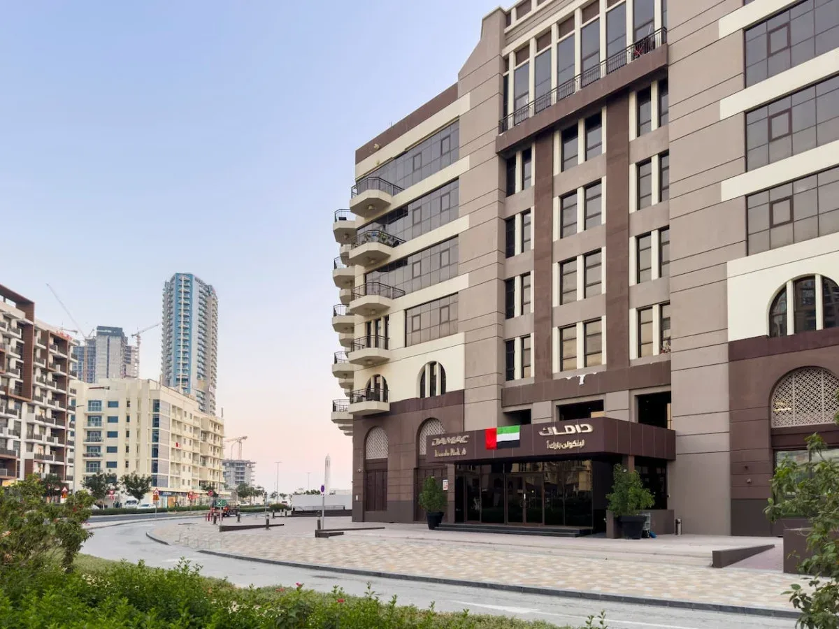 A modern multi-story building faces a paved street and landscaped greenery.