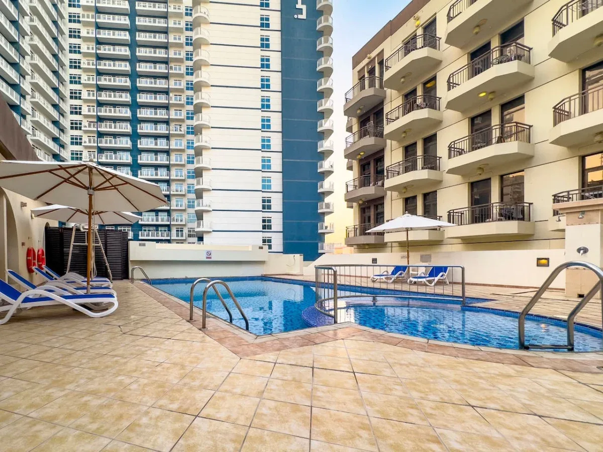 An outdoor swimming pool sits between tall apartment buildings, with lounge seating nearby.