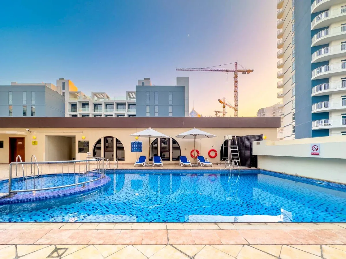 A rooftop swimming pool sits beside modern buildings, with umbrellas and loungers along the edge.