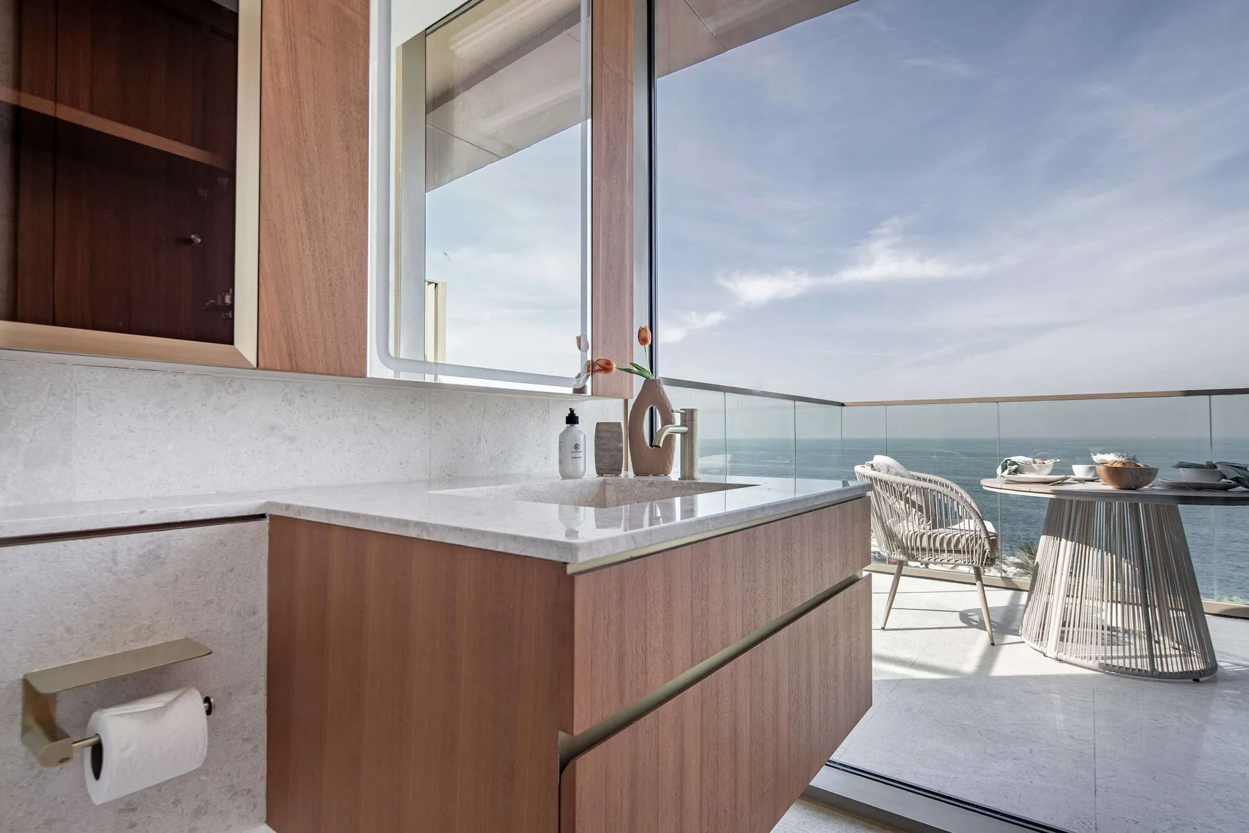 Un lavabo de baño da a un balcón con mesa y asientos frente al agua.