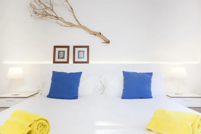A bright bedroom with white bedding, blue pillows, and yellow towels.