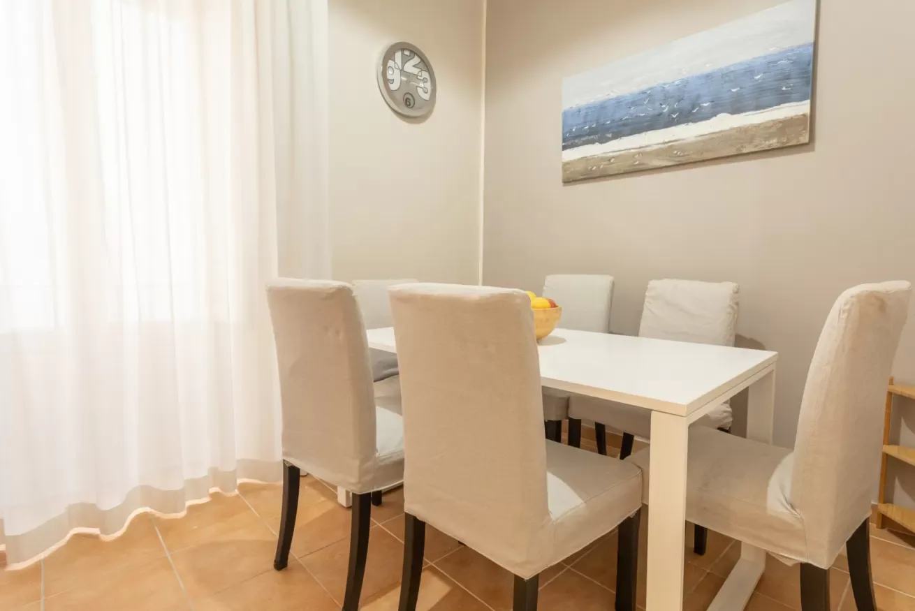 A dining area with a white table and upholstered seating beneath a coastal painting.