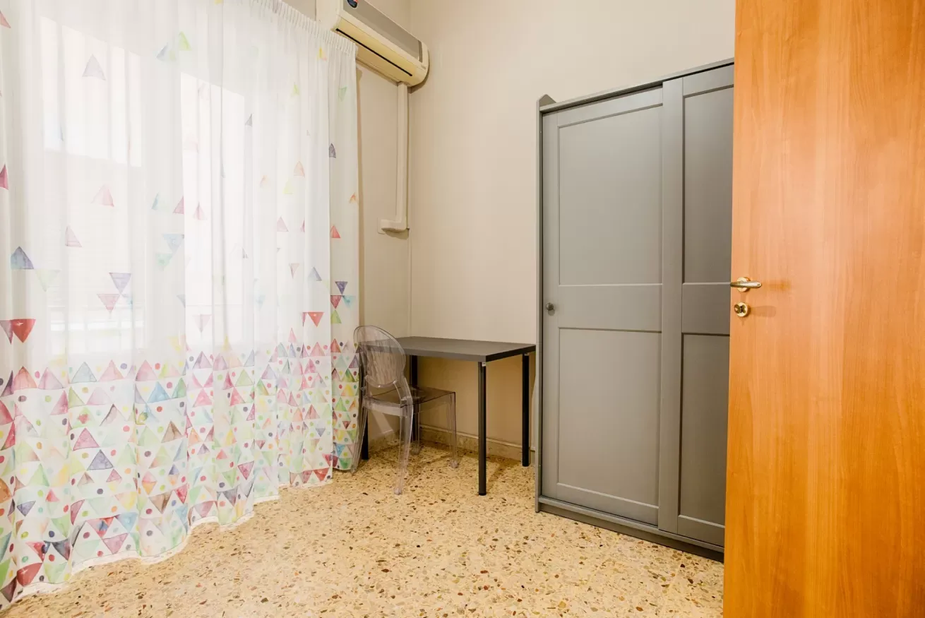 A small room with a table beside a curtained window and a gray wardrobe.