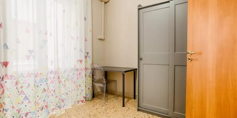 A room with a table beside a curtained window and a gray wardrobe.