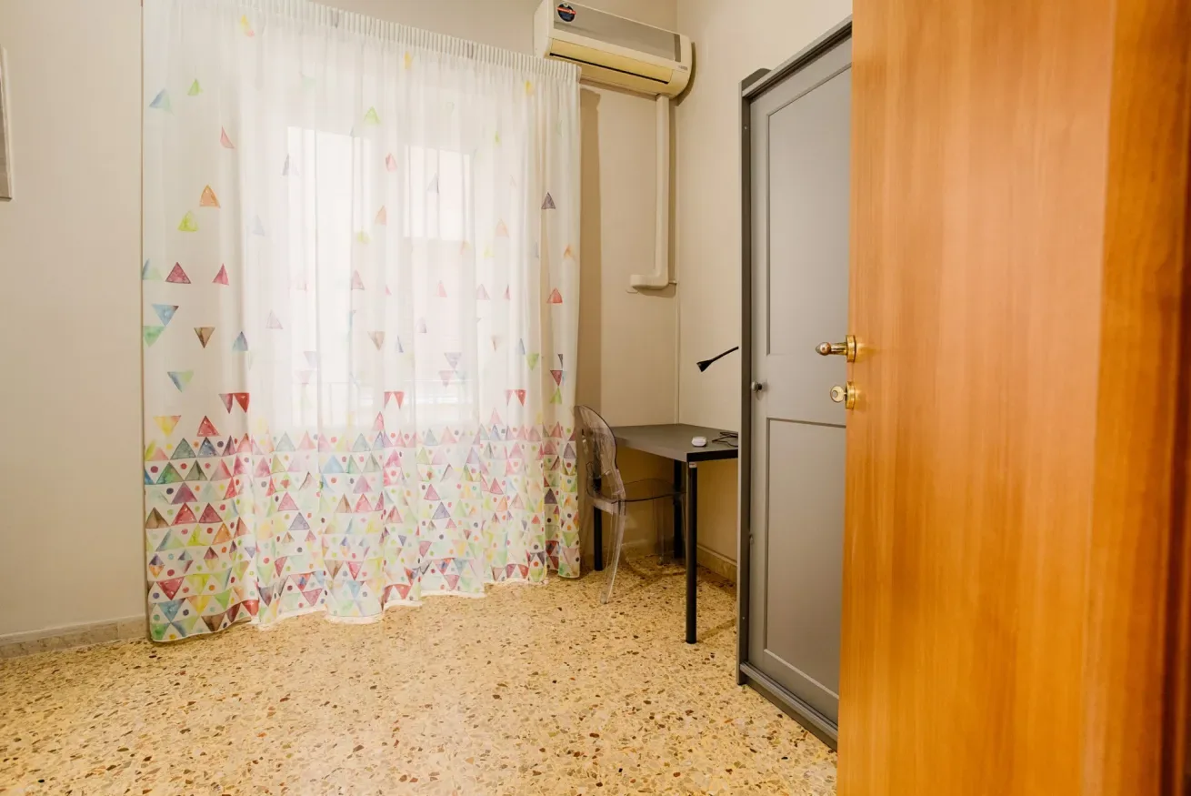 A room with patterned curtains, a small desk, and a door.
