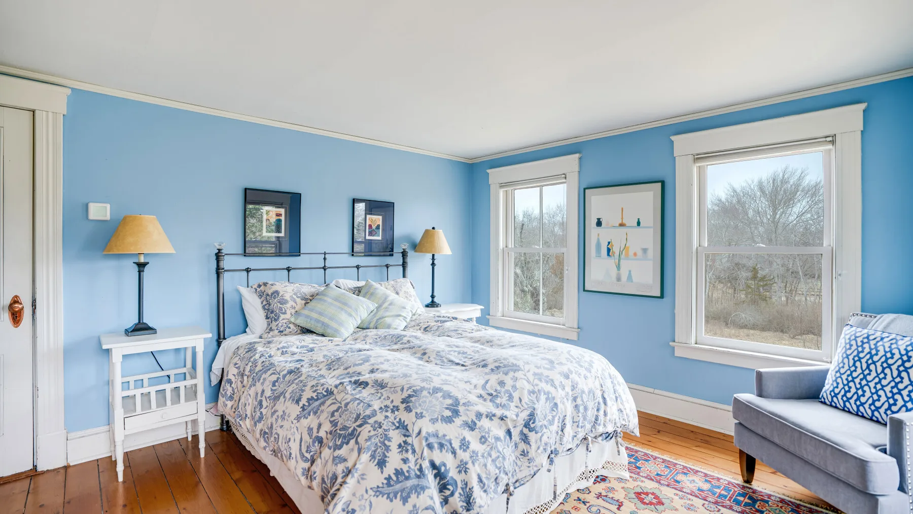 A bedroom with a bed, blue walls, and windows overlooking trees.