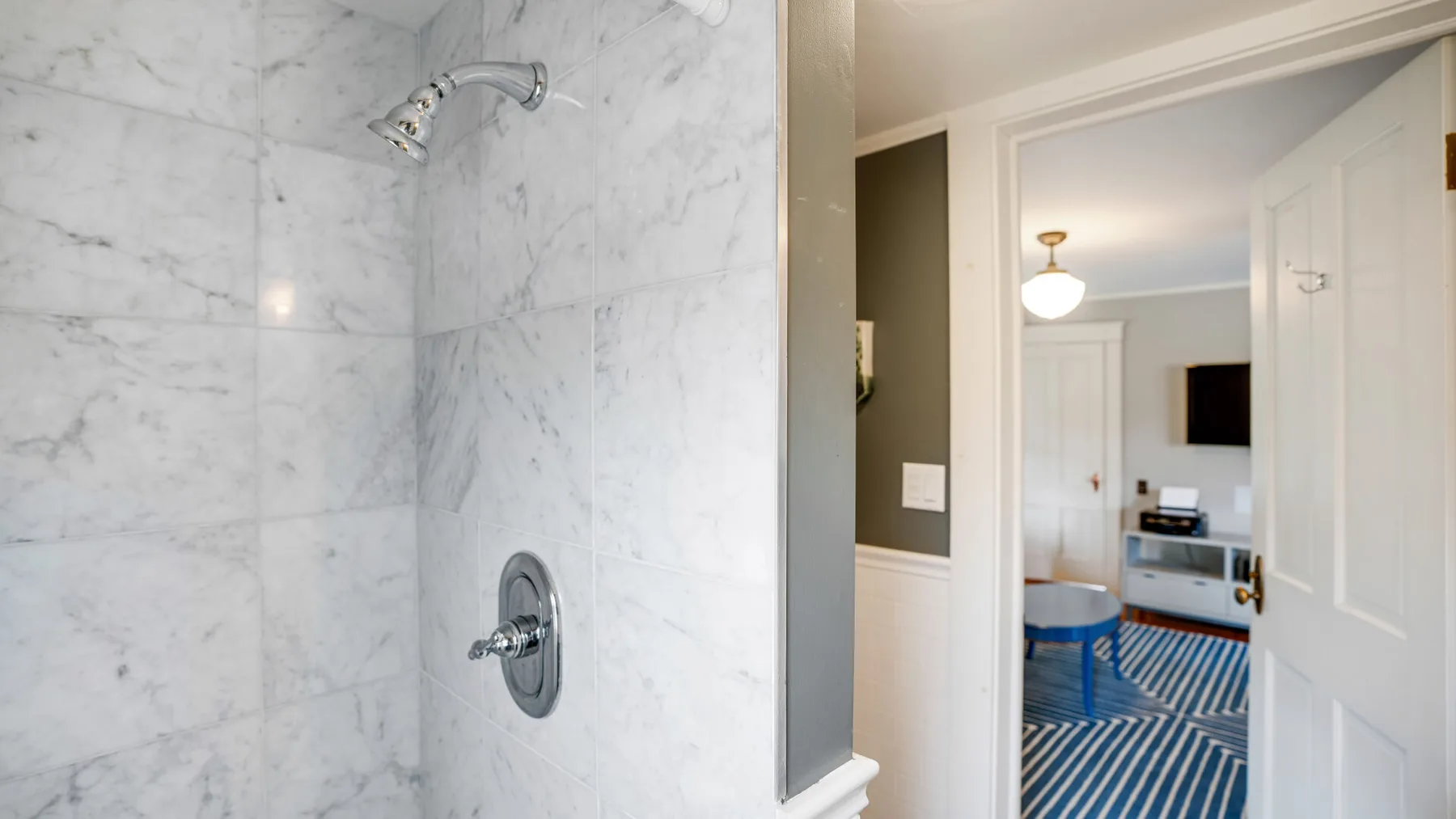 A marble-tiled shower opens onto a bedroom with a blue striped rug.