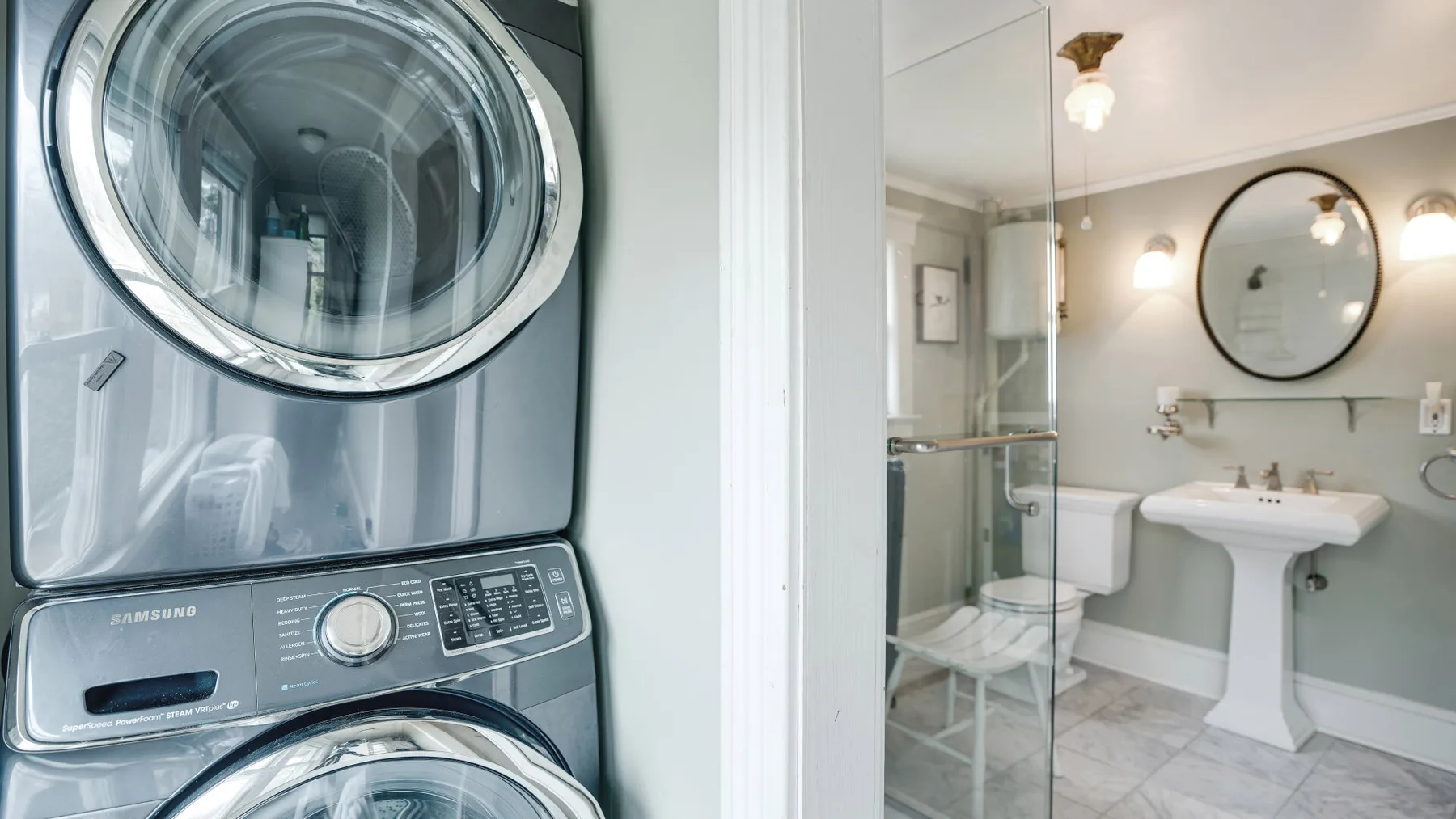 A bathroom with a pedestal sink and glass shower beside stacked laundry machines.
