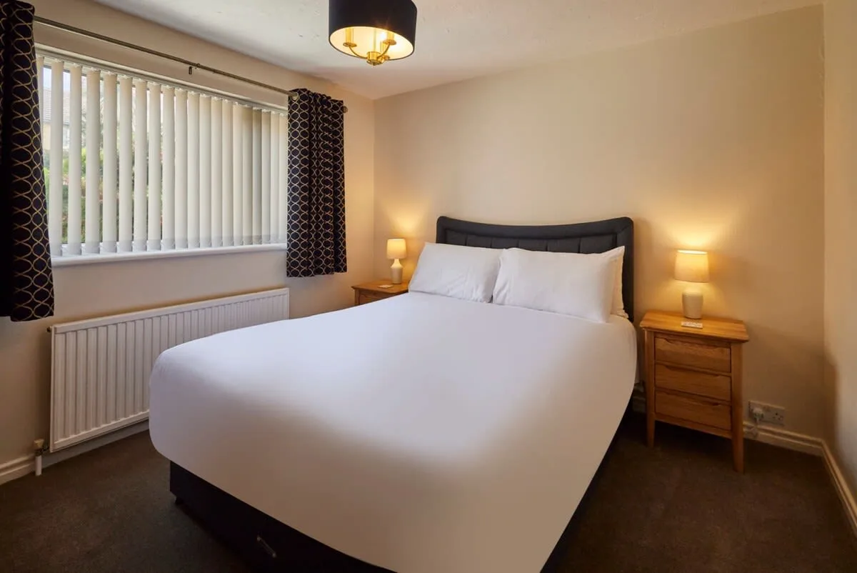 A bedroom with a neatly made bed, bedside tables, and a window with blinds.