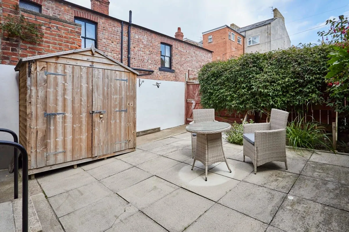 A paved courtyard with outdoor seating, a wooden shed, and a tall hedge.
