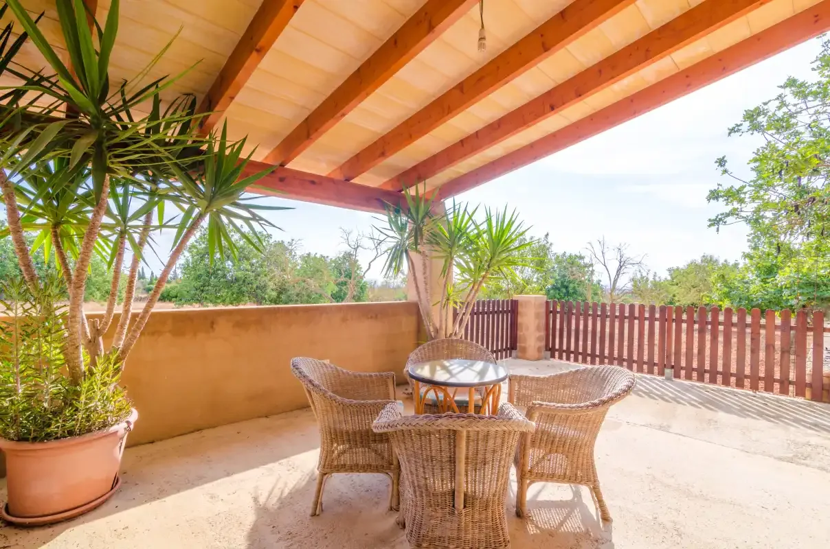 Un patio cubierto tiene una mesa redonda y asientos tejidos junto a una barandilla de madera.