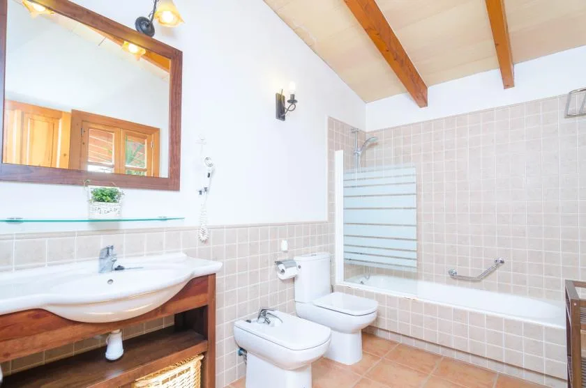 Un baño alicatado con bañera, inodoro y lavabo bajo vigas de madera vistas.