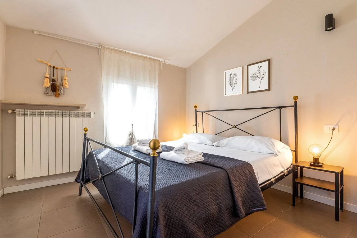 Un dormitorio con una cama de estructura metálica, ropa de cama blanca y una ventana con cortinas.