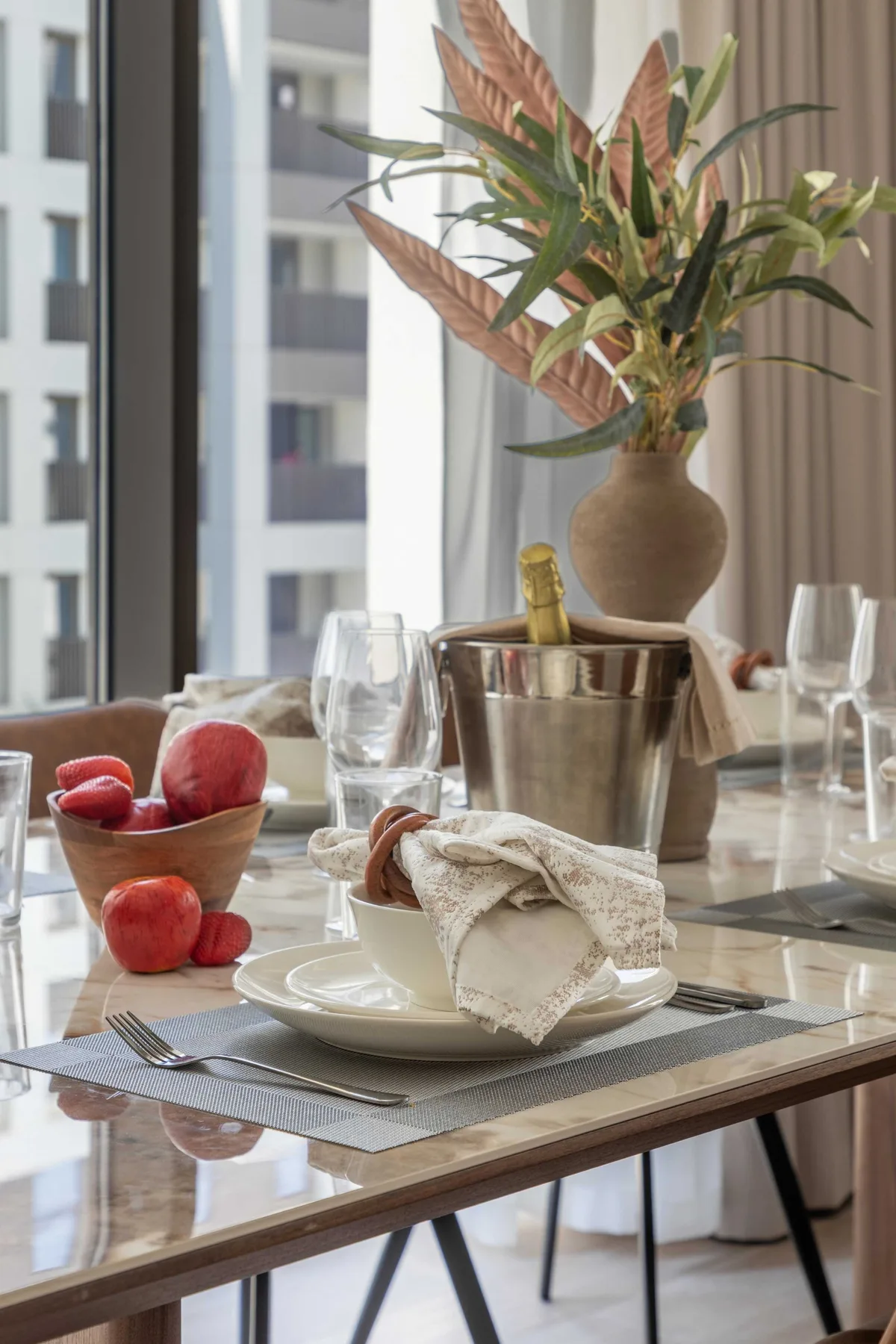 Una mesa de comedor está puesta con platos, copas, fruta y un jarrón con ramas.