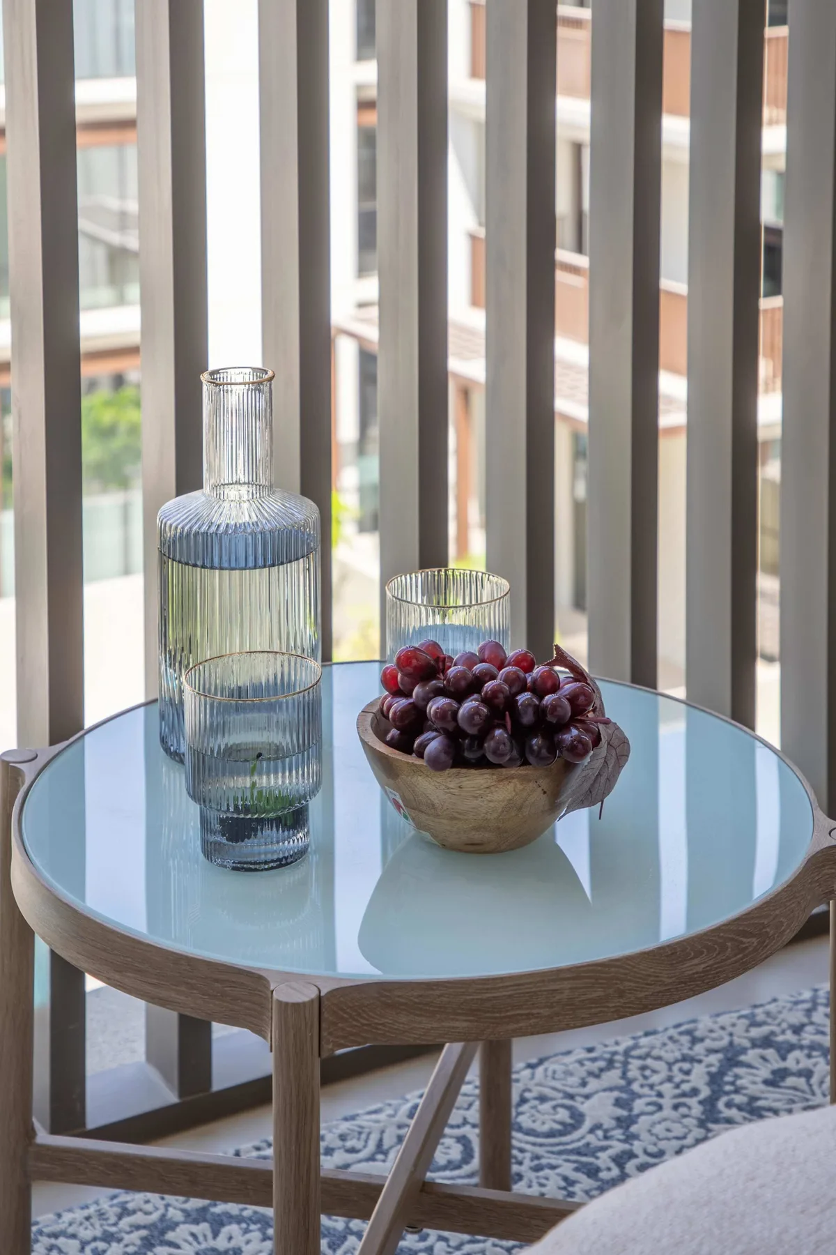 Una mesa redonda pequeña con un jarrón de vidrio y un cuenco de uvas está junto a ventanas altas.