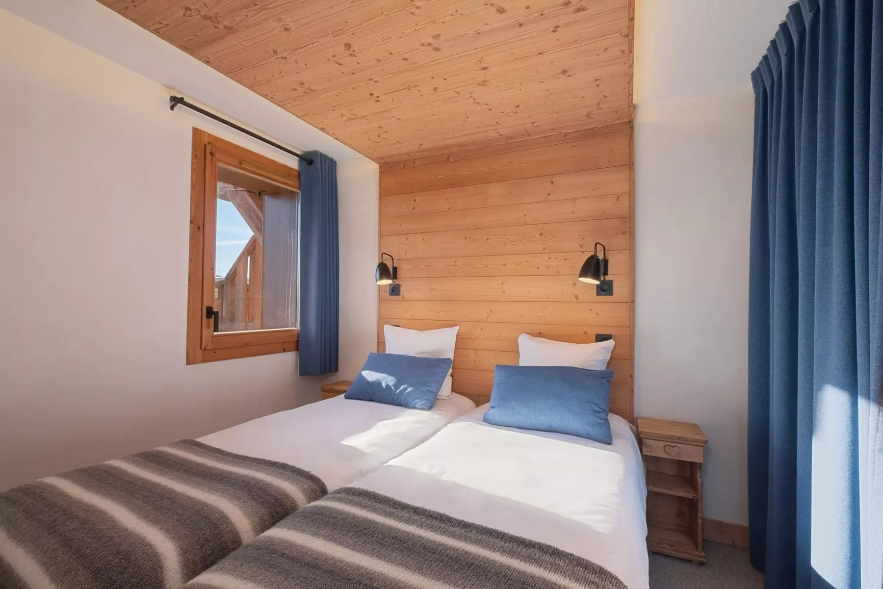 A bedroom with two beds, wooden wall paneling, and blue curtains.