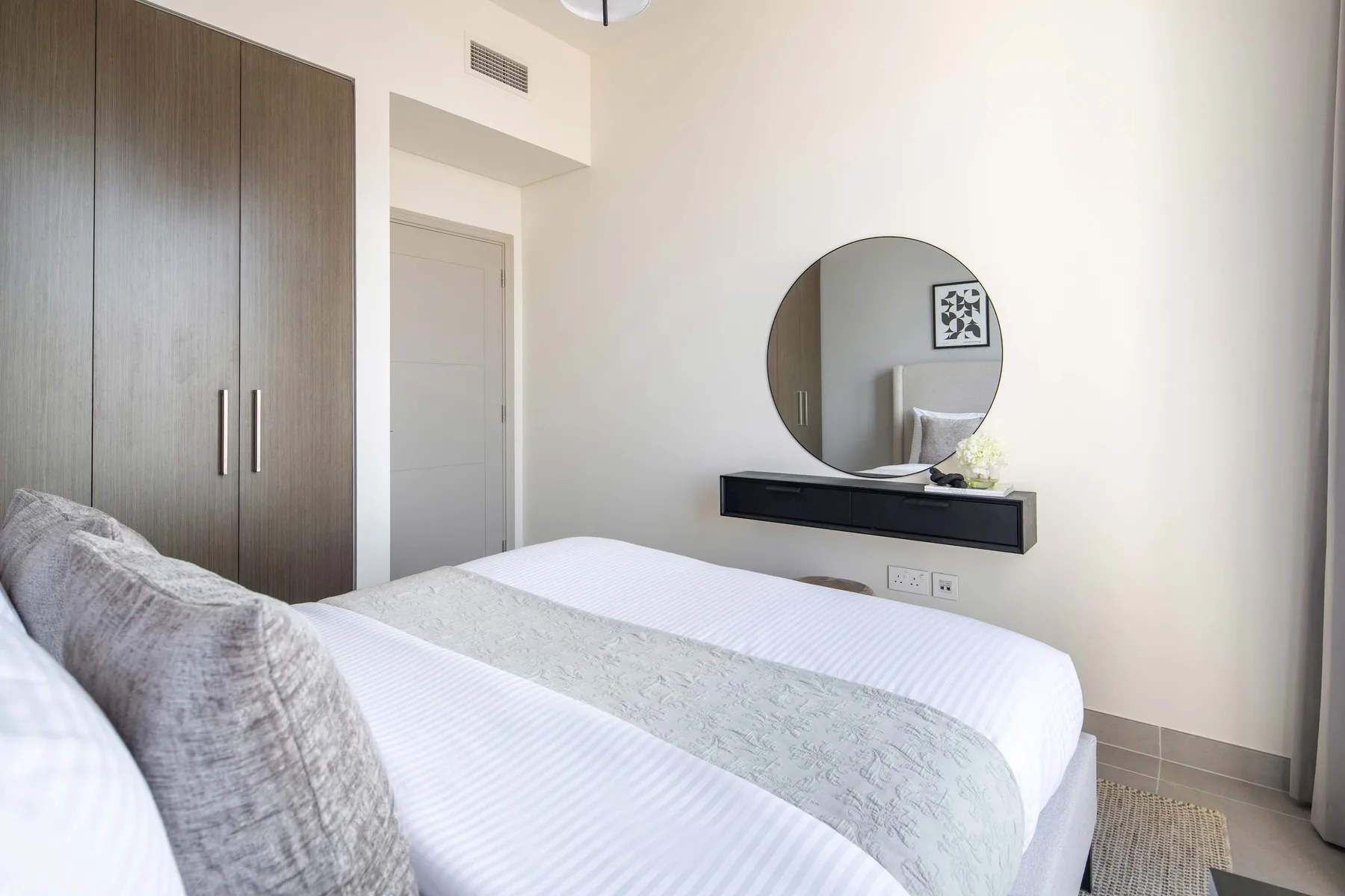A bright bedroom with a neatly made bed, dark wardrobes, and a round wall mirror.