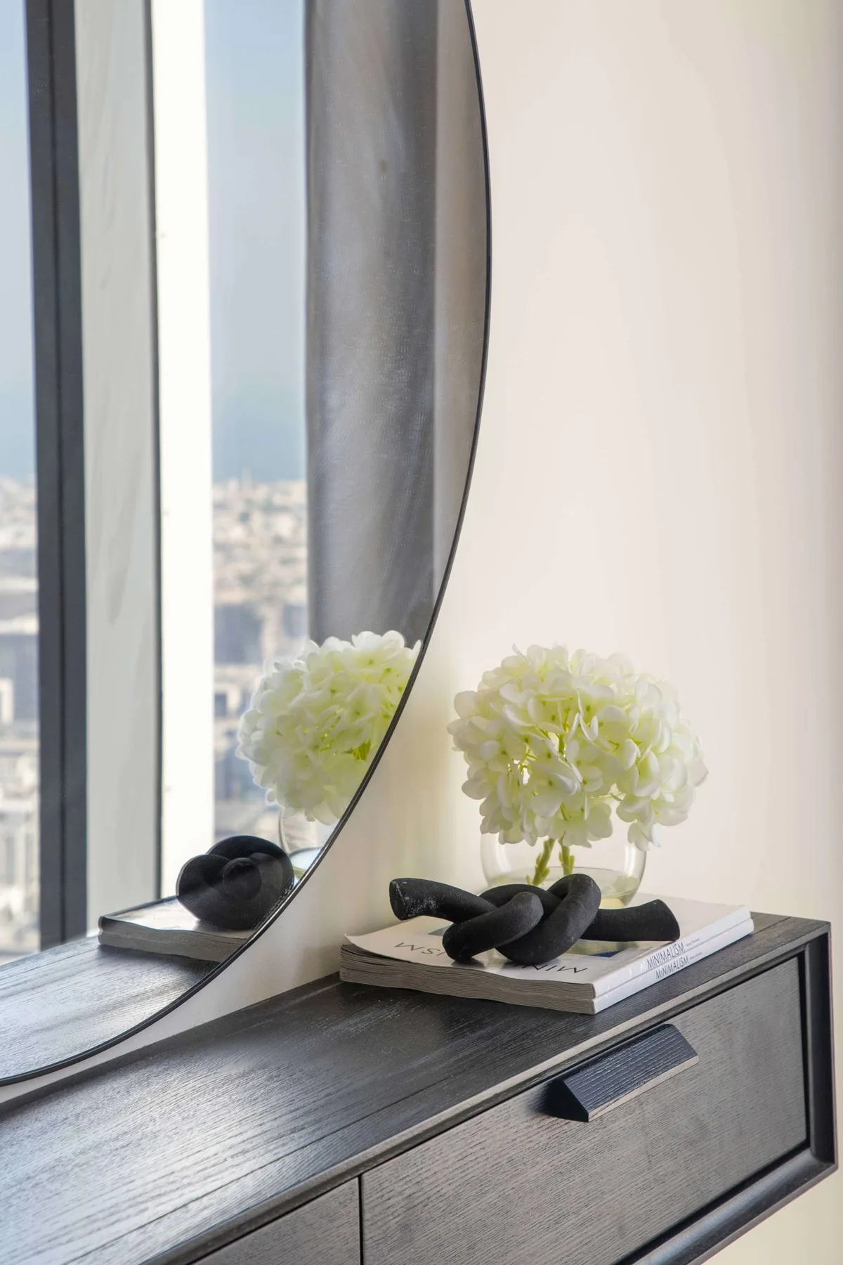 Bedroom dresser with a round mirror and white flowers beside a window.