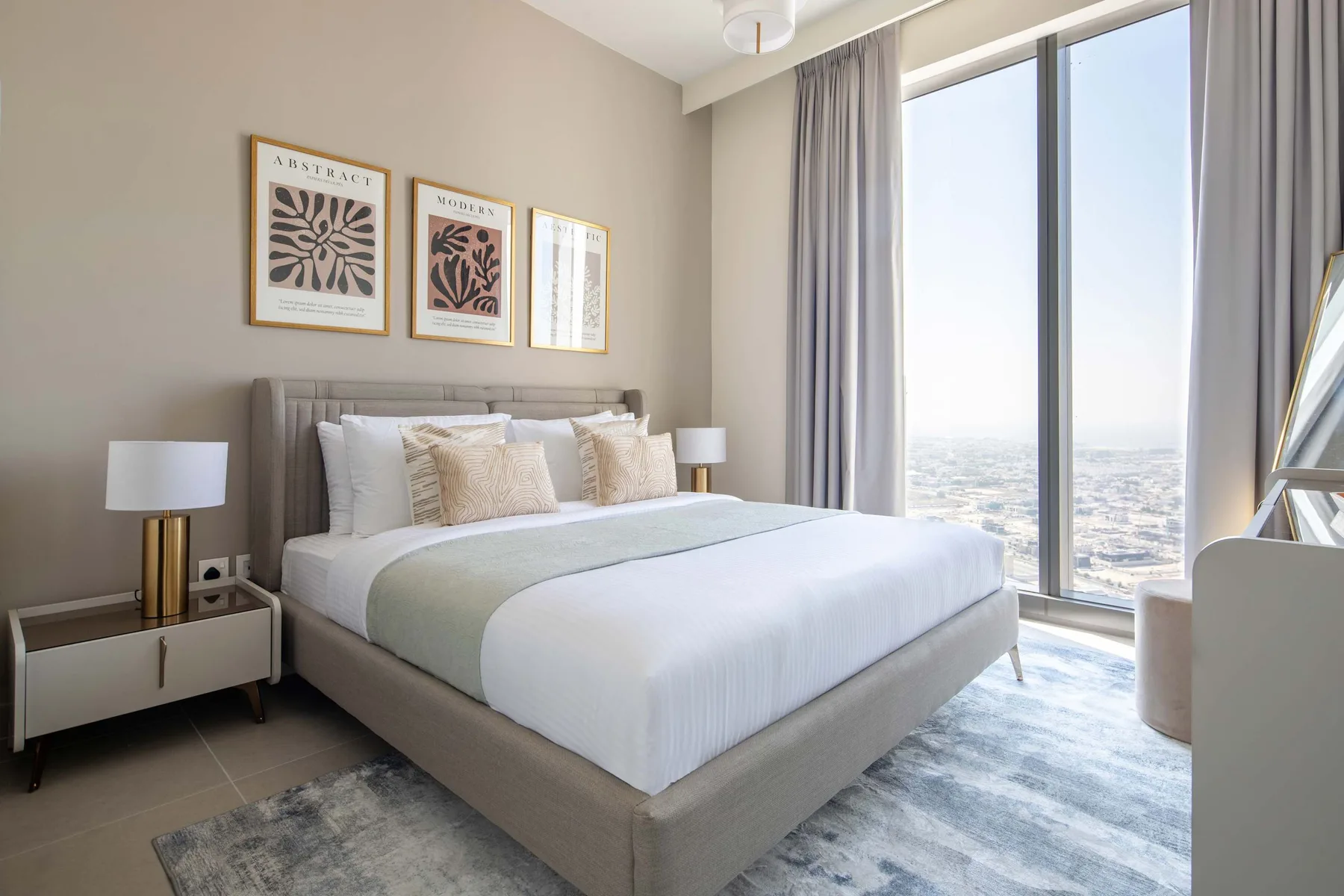 A bedroom with a neatly made bed beside tall windows overlooking the city.
