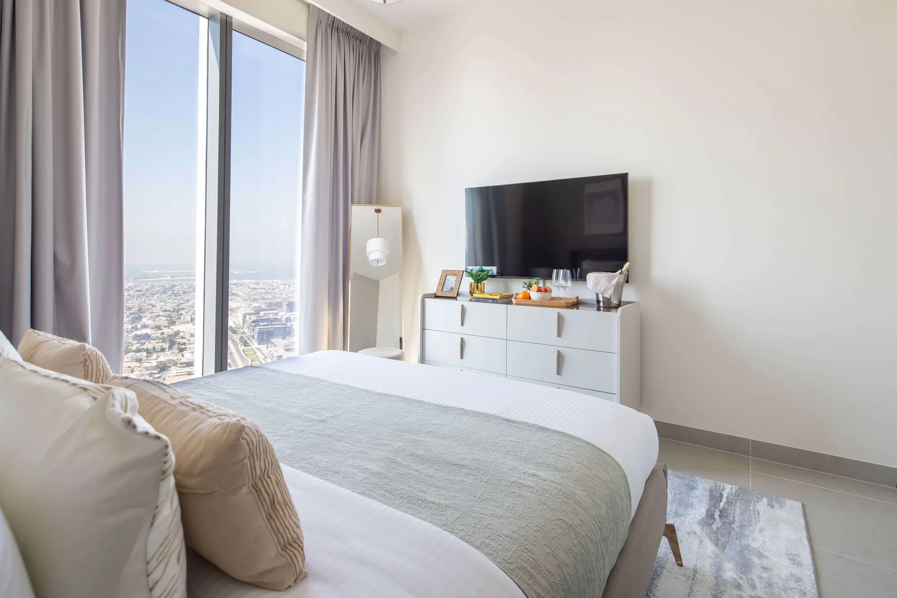 A bright bedroom with a bed beside tall windows and a dresser with a television.