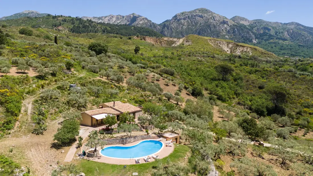 Una casa en la ladera y una piscina están rodeadas de matorrales verdes, con montañas al fondo.