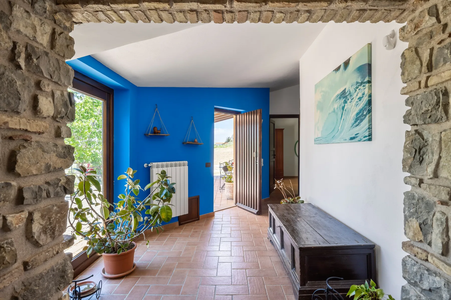 Un pasillo interior colorido da a un patio soleado, con paredes azules y una obra enmarcada.