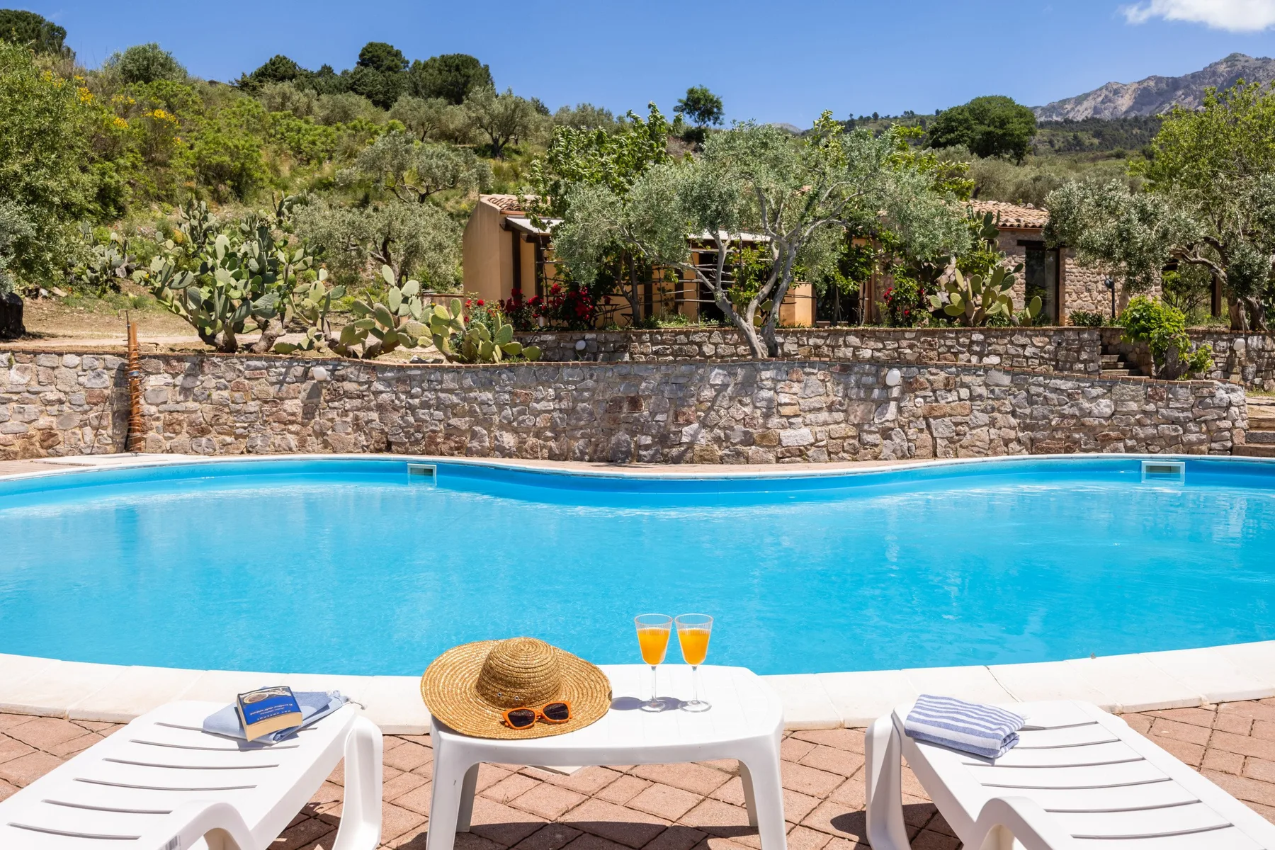 Una piscina está junto a un muro de piedra, con tumbonas y una mesa pequeña en primer plano.