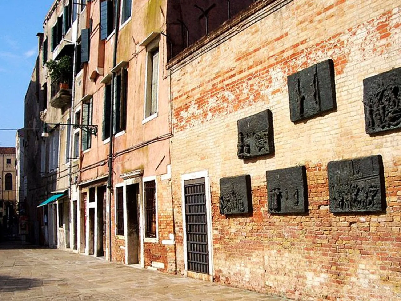 A sunlit stone-paved alley runs beside old brick buildings with dark wall reliefs.
