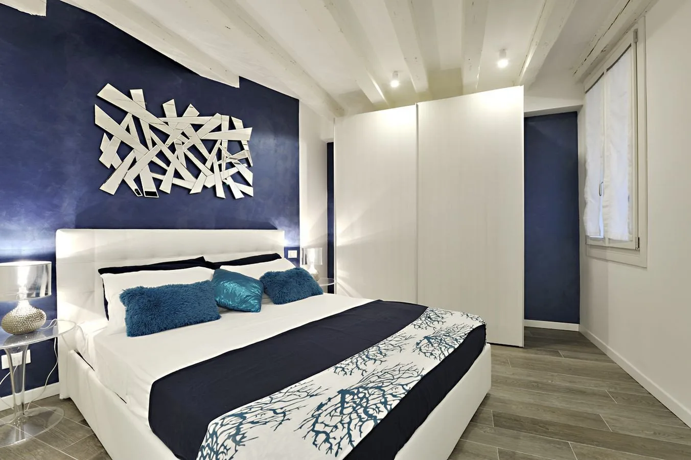 A bedroom with a white bed, dark blue accent wall, and abstract wall art.