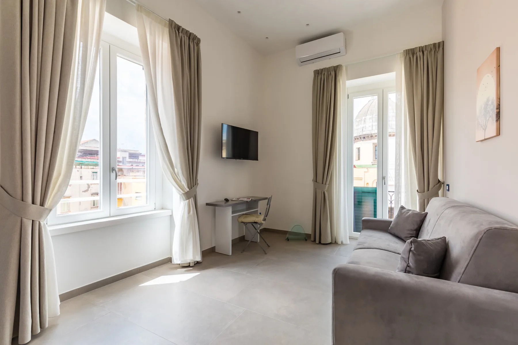 Una sala luminosa con asientos, un televisor y ventanas altas con cortinas.