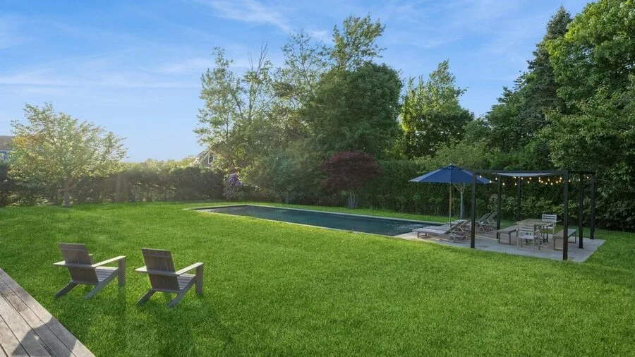 A grassy backyard with a long pool, outdoor seating, and a shaded patio.