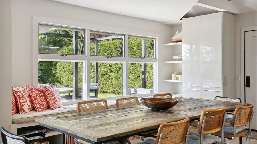 A bright dining area with a long table beside large windows.