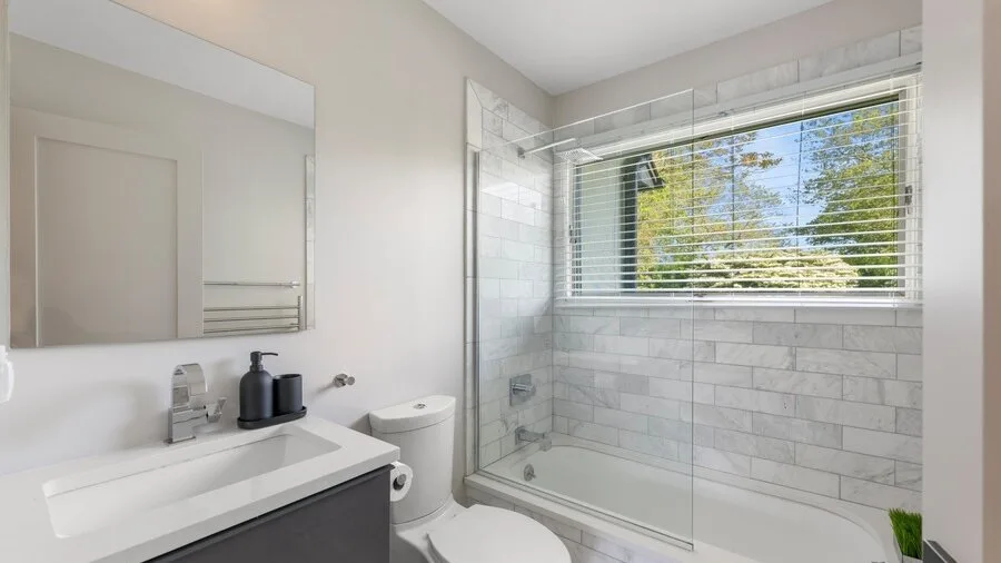 A bright bathroom with a sink, toilet, and a shower beside a window.