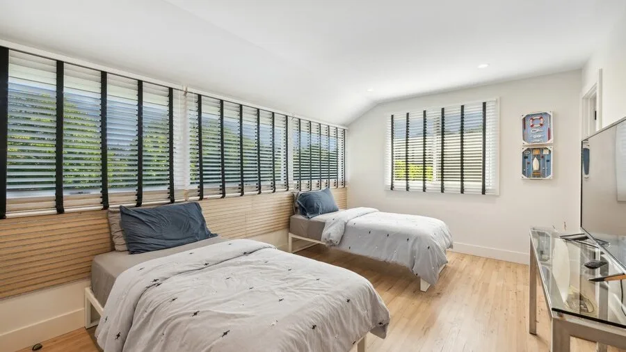A bright bedroom with two beds beside wide windows fitted with blinds.