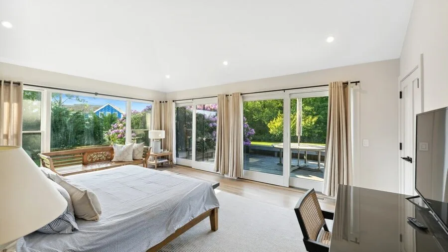 A bright bedroom with a bed beside large windows overlooking greenery.